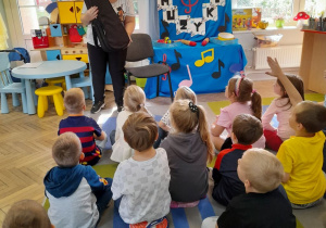 Dzieci siedzą na dywanie, a przed nimi stoi Pani, która trzyma futerał z instrumentem. Dzieci snują domysły jaki instrument znajduje się w futerale. W tle dekoracja z okazji Dnia Muzyki.