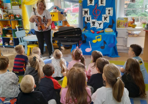 Dzieci siedzą na dywanie, a mama Bartka prezentuje skrzypce.