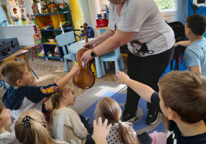 Dzieci siedzą na dywanie i dotykają drewnianej części skrzypiec, które trzyma rodzic.