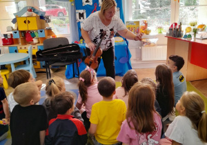 Dzieci siedzą na dywanie, a przed nimi Pani prezentuje kalafonię do nacierania włosia smyczka.