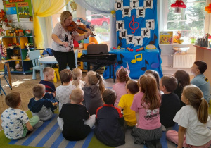 Dzieci siedzą na dywanie, a przed nimi mama Bartka gra na skrzypcach.