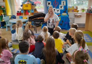 Dzieci razem z Panią siedzą na dywanie. Mama Bartka pokazuje przedszkolakom gdzie znajdują się struny głosowe.