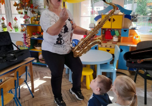 Pani prezentuje dzieciom saksofon. W tle okna oraz kąciki zainteresowań: domowy i przyrodniczy.