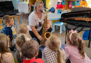 Mama Bartka przykucnęła obok dzieci na dywanie i gra na saksofonie.