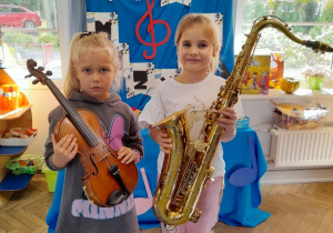 Dwie dziewczynki stoją obok siebie. Tosia trzyma skrzypce, a Marysia saksofon. W tle tablica z napisem Dzień Muzyki.