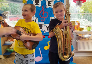 Dwóch chłopców stoi obok siebie. Antek trzyma skrzypce, a Marcel saksofon.