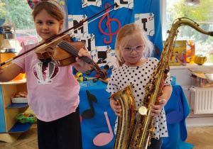 Dwie dziewczynki stoją z instrumentami obok siebie. Nadia trzyma skrzypce, a Zuzia saksofon.