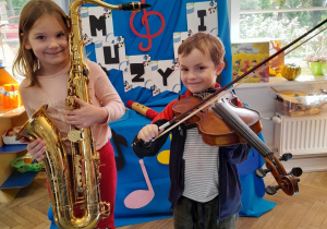 Dwoje dzieci pozuje do zdjęcia na tle dekoracji z okazji Dnia Muzyki. Weronika trzyma saksofon, a Bartek skrzypce.