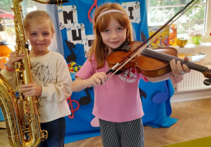 Dwie dziewczynki stoją obok siebie z instrumentami muzycznymi. Nikola trzyma saksofon, a Emma skrzypce.