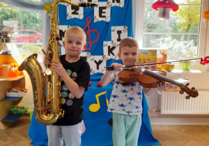 Dwóch chłopców stoi na tle dekoracji z okazji Dnia Muzyki. Fabian trzyma saksofon, a Mikołaj skrzypce.