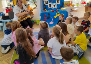 Przedszkolaki siedzą na dywanie, a Pani siedzi na krześle trzymając saksofon i opowiada dzieciom o instrumencie. W tle okna oraz dekoracja z okazji Dnia Muzyki.