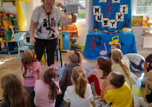 Dzieci siedzą na dywanie, a Pani pokazuje dzieciom lirkę do trzymania nut.