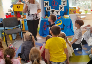 Dzieci siedzą na dywanie, a mama Bartka pokazuje kartę pracy z zakodowanym instrumentem.