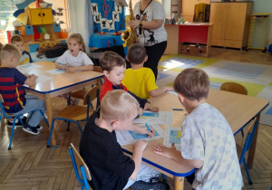 Dzieci siedzą przy stolikach i kolorują obrazki, a Pani gra na saksofonie.