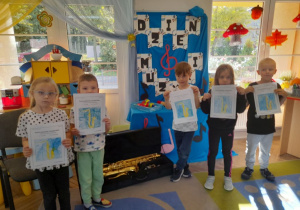 Grupka dzieci prezentuje wykonane prace. W tle dekoracja z napisem Dzień Muzyki, kącik domowy, a na dywanie leży saksofon w futerale.