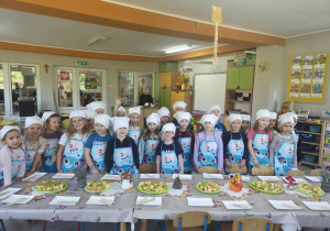 1.Przedszkolaki ubrane w fartuszki i białe chustki na głowach stoją przy stole zastawionym owocami