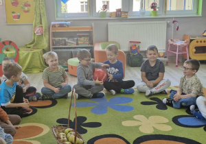 6.Dzieci siedzą na dywanie podając sobie czerwoną piłkę symbolizującą jabłko. Przed nimi leży kosz z jabłkami