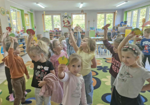 8.Przedszkolaki stoją na dywanie i unoszą w dłoniach papierowe kolorowe jabłuszka