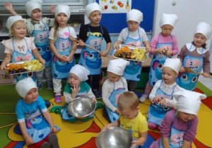 Dzieci z grupy Pszczółek w fartuszkach i białych chusteczkach pozują do zdjęcia. Maja i Olaf trzymają tace z owocami.