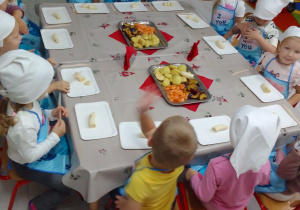 Dzieci w fartuszkach i białych chusteczkach na głowach siedzą przy stolikach, na których są tace, noże i owoce oraz serwetki.