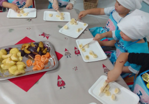 Dzieci w fartuszkach i białych chusteczkach samodzielnie kroją owoce na sałatkę.