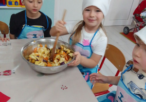 Kornelia miesza drewnianą łyżką wszystkie składniki sałatki owocowej. Obok Leon i Michał.