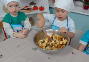 Olaf miesza łyżką kawałki owoców znajdujące się w misce. Obok przy stole siedzi Olek.