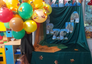 Dekoracja z okazji Dnia Drzewa - na zielonym materiale przypięty jest napis Dzień Drzewa oraz listki w jesiennych barwach. Obok stoi drzewo z balonów.