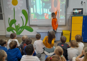 Dzieci siedzą na dywanie, a Mikołaj pokazuje na tablicy multimedialnej dziuplę w drzewie.