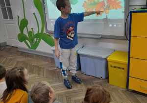 Grupka dzieci siedzi na dywanie, a Antoś wskazuje na tablicy multimedialnej owoce i liście dębu.