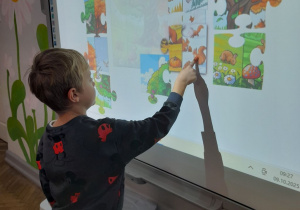 Hubert stoi przed tablicą multimedialną i układa puzzle "Drzewo w czterech porach roku".