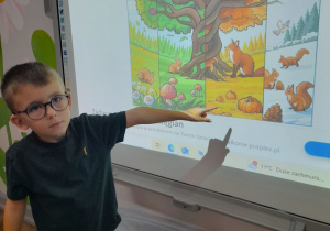 Na tablicy multimedialnej wyświetlone jest drzewo w czterech porach roku. Mikołaj wskazuje fragment jesiennego drzewa.