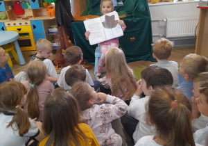 Dzieci siedzą na dywanie, a przed nimi na krzesełku Marysia. Dziewczynka pokazuje w albumie zasuszony liść tulipanowca.