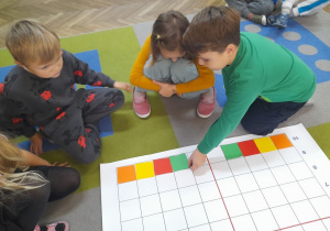Julek układa na dole maty do kodowania kolorowe kartki według podanej instrukcji. Obok siedzi dwoje dzieci.