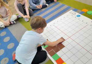 Na dywanie leży mata do kodowania. Staś układa na macie pień drzewa z brązowych kartonów. Obok siedzi grupka rówieśników.