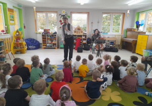 Dzieci siedzą na dywanie, a przed nimi stoi Pan prowadzący audycję muzyczną. Obok siedzi Pan z wiolonczelą. W tle kąciki zabaw.