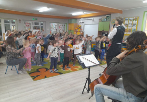 Dzieci stoją na dywanie i rytmicznie poruszają rękami przy dźwiękach wiolonczeli.