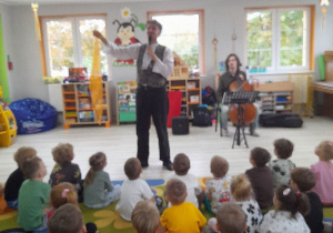 Pan prowadzący audycję stoi na środku sceny i opowiada dzieciom bajkę o czarodziejce Wioli.