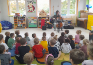 Dzieci siedzą na dywanie, a na środku sceny siedzi Pan, który gra na wiolonczeli.