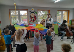 Pan gra na wiolonczeli, a dzieci podnoszą do góry chustę animacyjną.