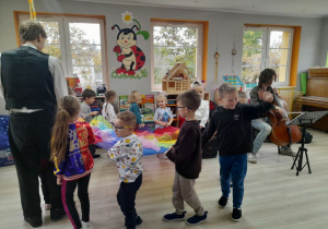 Dzieci razem z Panem prowadzącym audycję trzymają chustę animacyjną i poruszają się przy muzyce. Obok Pan gra na wiolonczeli.