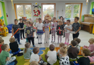 Dzieci siedzą na dywanie, a przed nimi grupa rówieśników trzyma instrumenty perkusyjne: rurki, tamburyno.