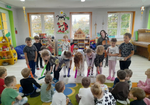 Dzieci siedzą na dywanie, a przed nimi grupa rówieśników gra na rurkach uderzając instrumentem rytmicznie o podłogę.