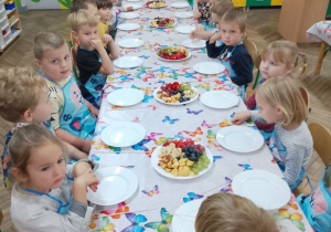 3.Grupa „Motylków” ubrana w fartuszki siedzi przy stole na którym stoją talerze z owocami.