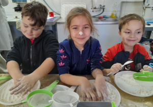 Troje dzieci stoi obok siebie przy stole podczas zabawy mąką. Amanda ma stempelek z mąki na nosie.