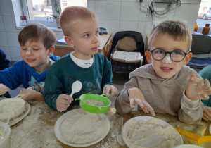 Trzech chłopców stoi obok siebie podczas zabawy z mąką. Hubert trzyma sitko, a Staś i Bartek mają stempelki z mąki na nosach.