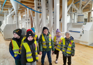 Grupka dzieci stoi obok siebie podczas zwiedzania młyna. W tle maszyny do mielenia zbóż.