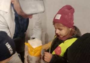 Obok pracownika młyna stoi grupka dzieci. Pan pokazuje przedszkolakom kaszę mannę.