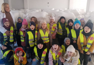 Dzieci podczas zwiedzania magazynu pozują do zdjęcia z Panią właścicielką. W tle worki z mąką.