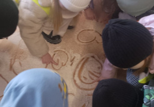 Grupka dzieci przykucnęła i rysuje wzory w mące rozsypanej na podłodze.